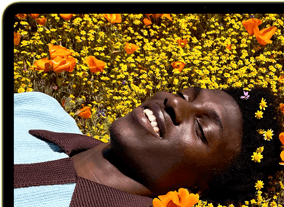 MacBook Neo, citrus colour, screen shows a smiling person lying in a sunny field of flowers