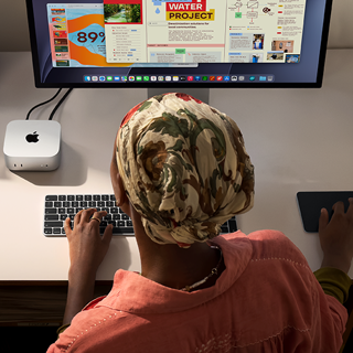 A person typing at a desk with a Mac mini, Studio Display connected, Magic Keyboard, Magic Trackpad, various items